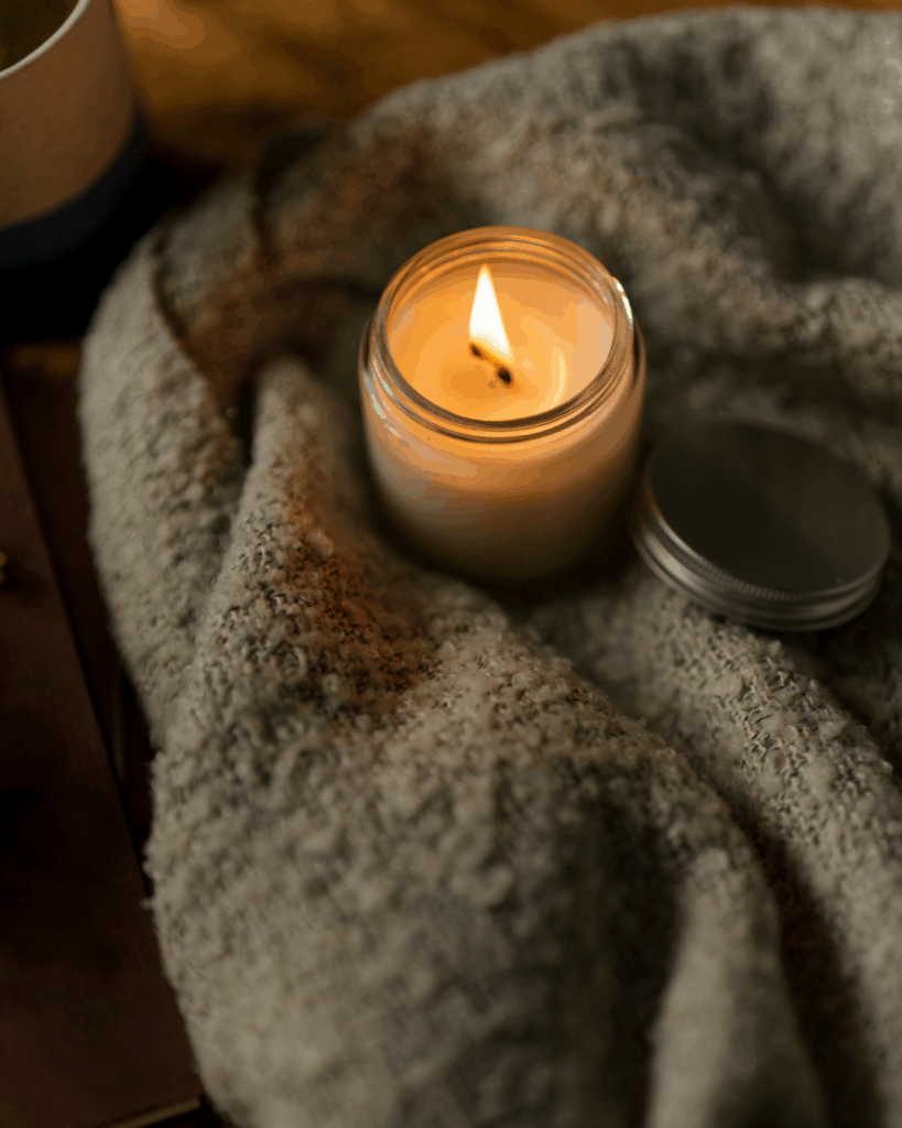 Зажжённая свеча стоит на мягком сером пледе — тепло, свет и атмосфера домашнего уюта.
